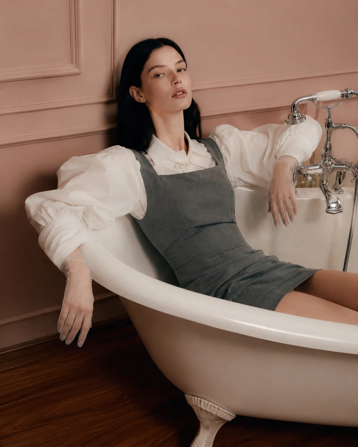 A woman in a sculptural white blouse and grey dress reclines in a freestanding bath against a muted pink interior.