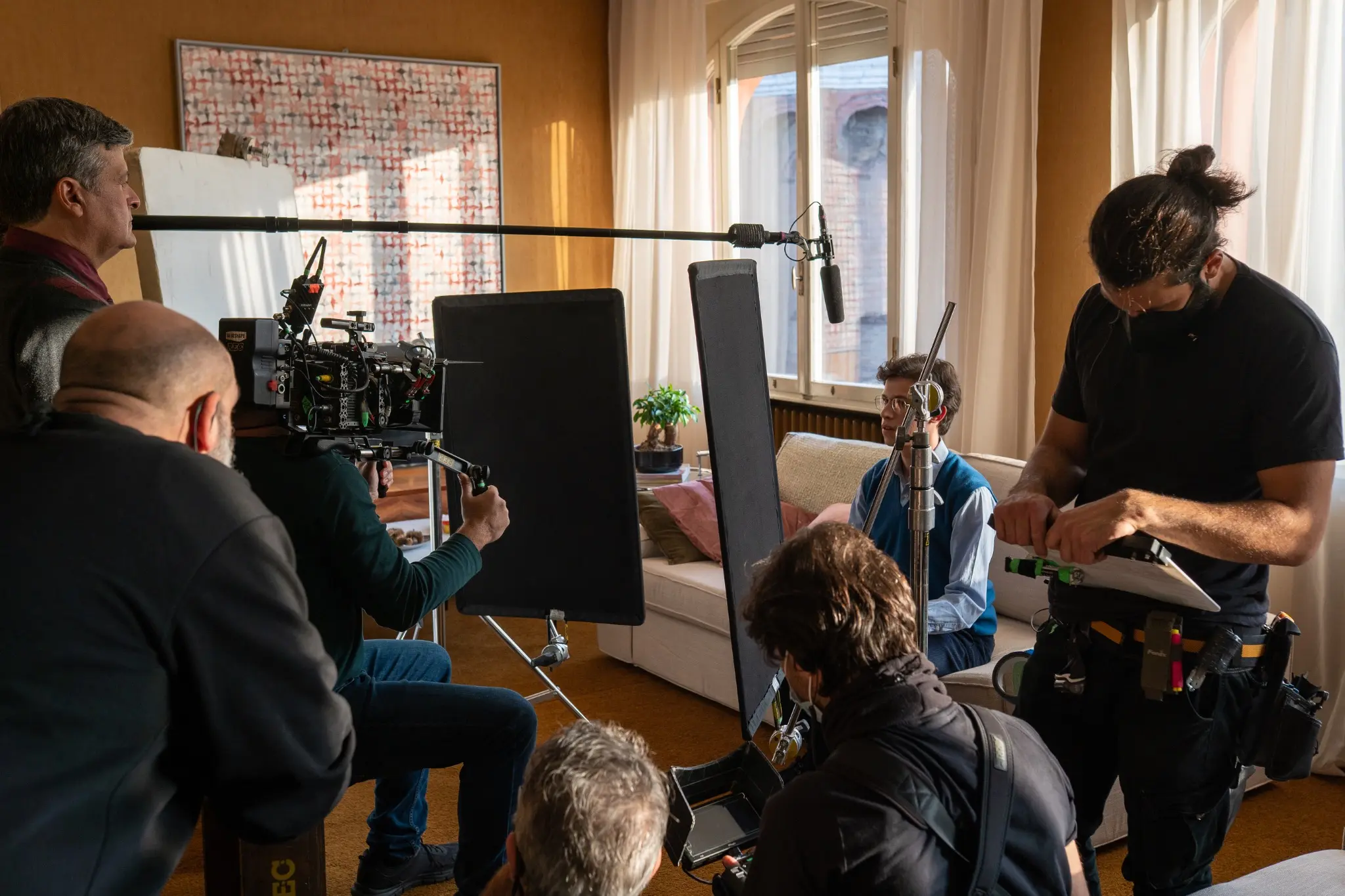 Camera, lighting and sound crew work around a performer in a daylight interior.