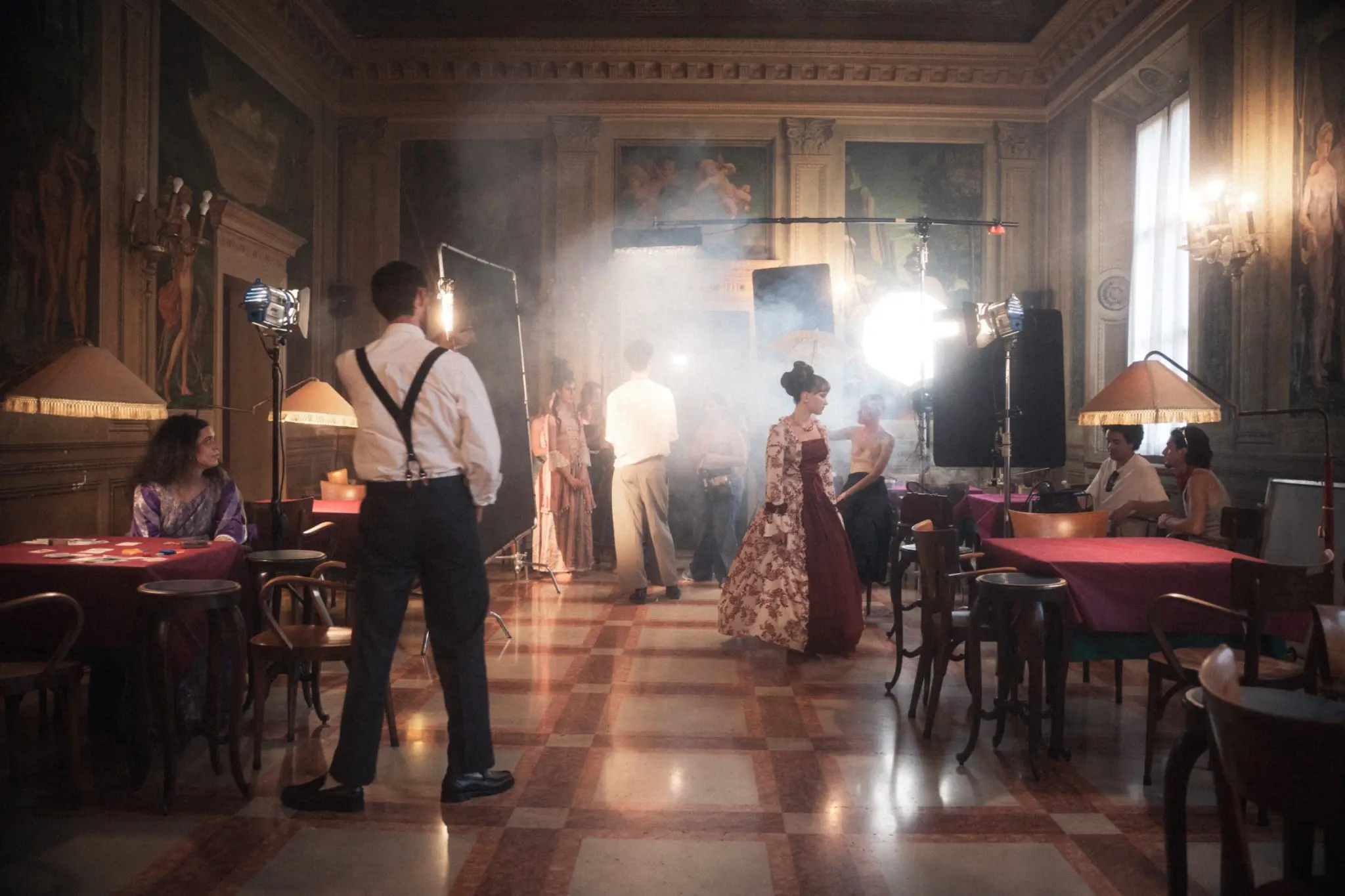 A film crew works inside a period interior filled with haze and practical light.