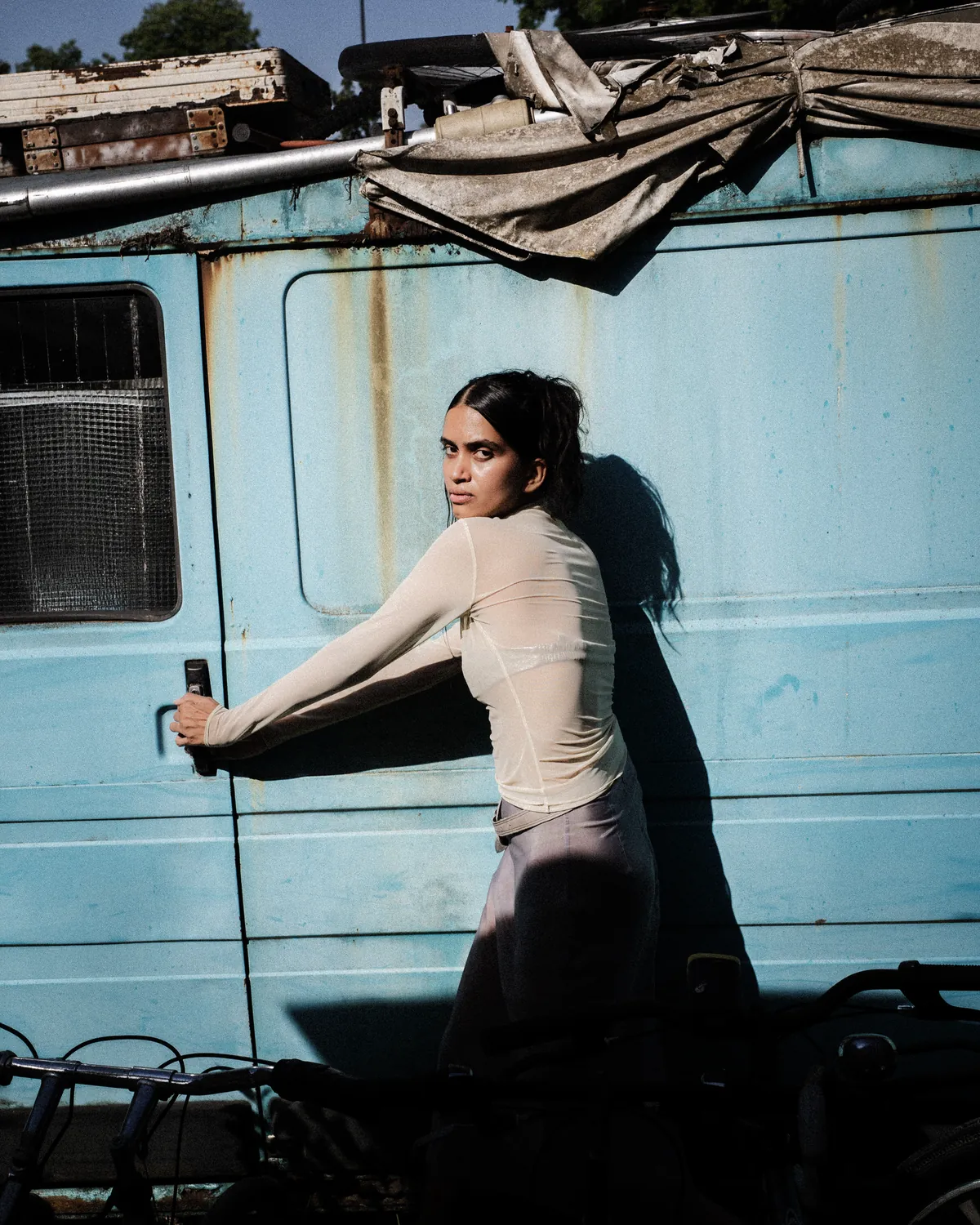 A model in a translucent top sits against a weathered blue vehicle in hard sunlight.