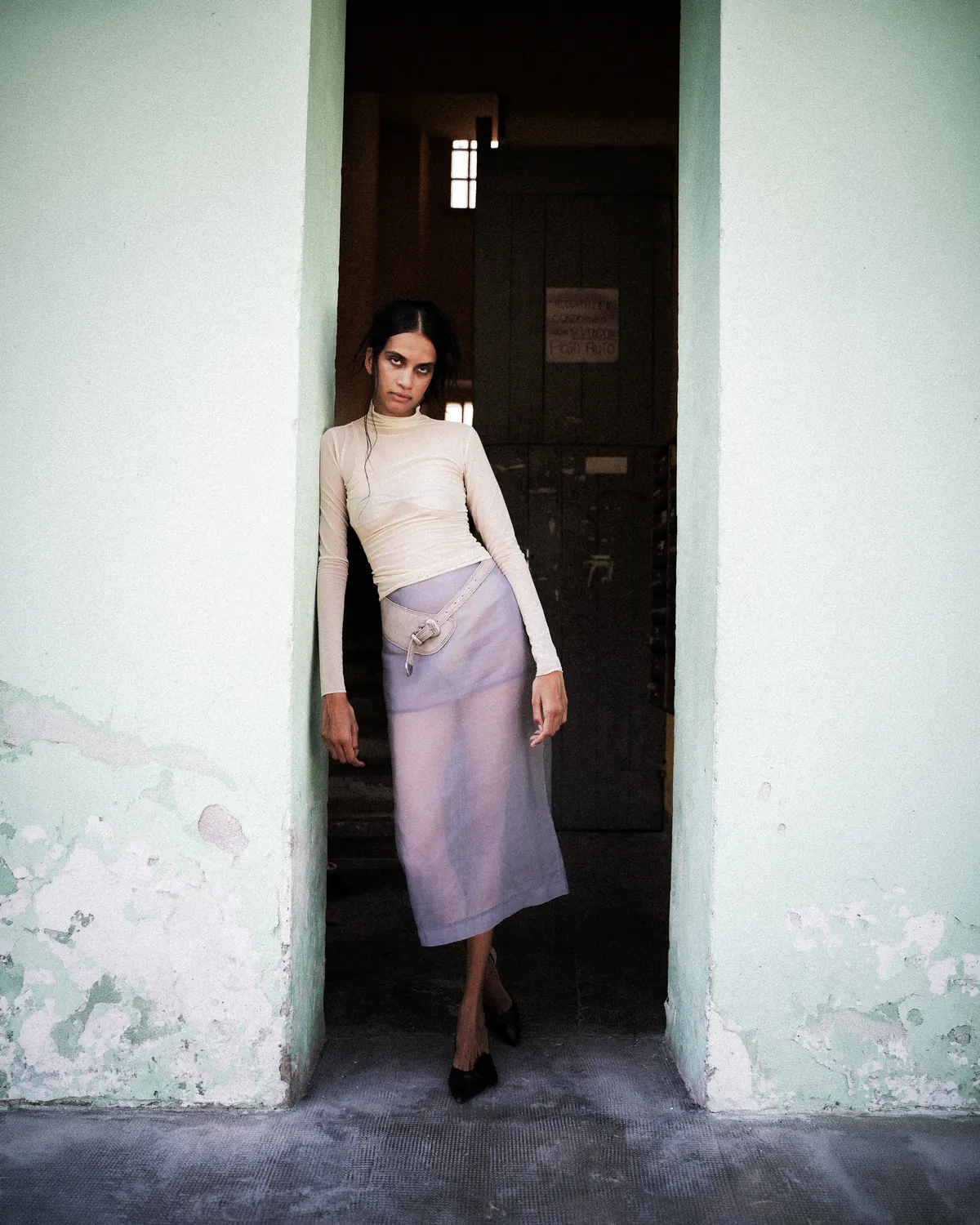 A model bends through the frame of a dark industrial doorway in strong daylight.